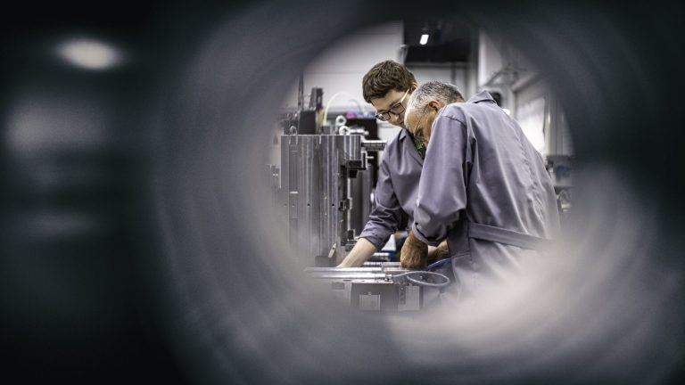 Two engineers in grey overalls leaning over machinery in a workshop, seen through a circular opening