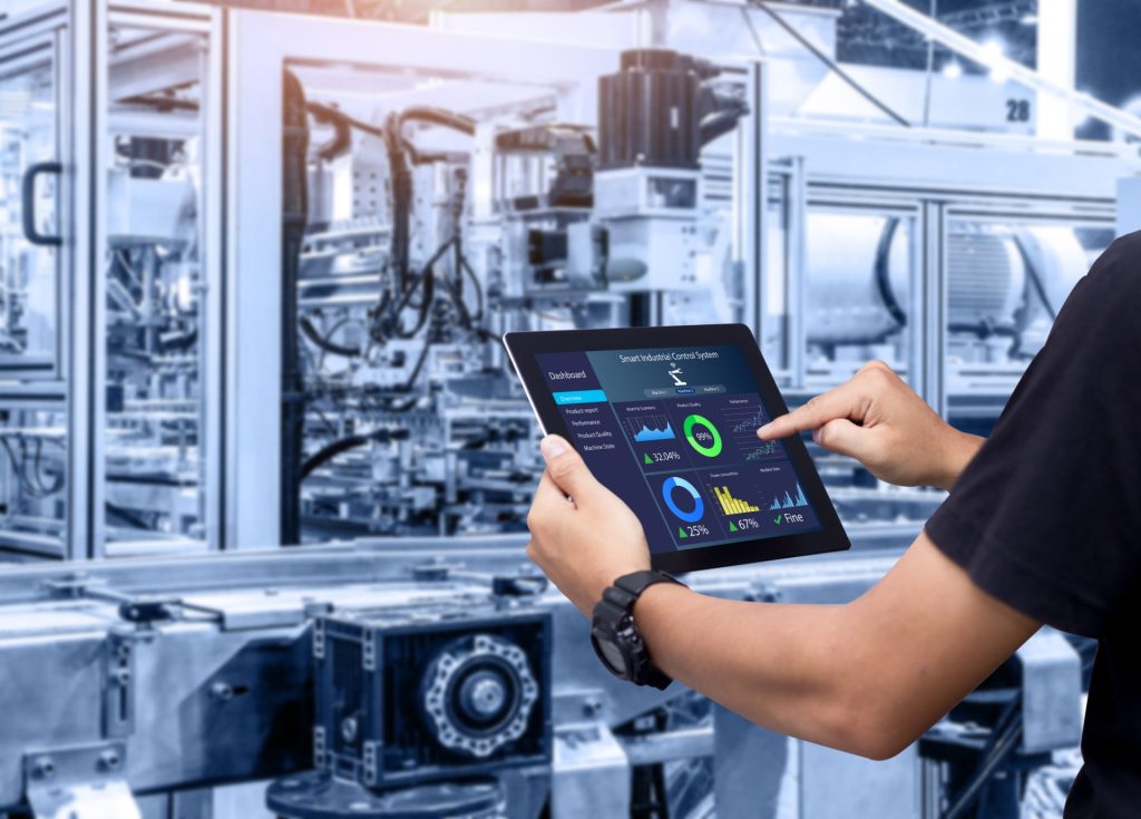 Engineer using a tablet showing an industrial control dashboard in front of automated manufacturing machinery