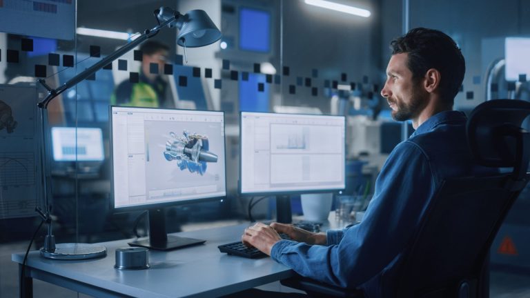 Engineer at a desk using two monitors, one showing a 3D model of a turbine, in an industrial control room
