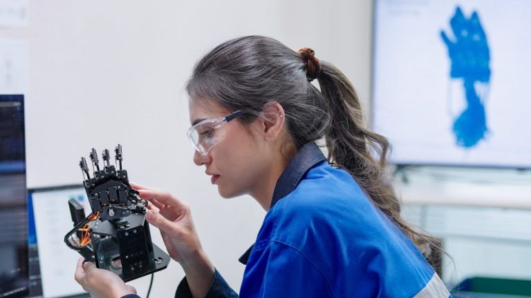 Engineer in safety glasses and blue overalls examining a robotic hand prototype in a lab beside computer screens