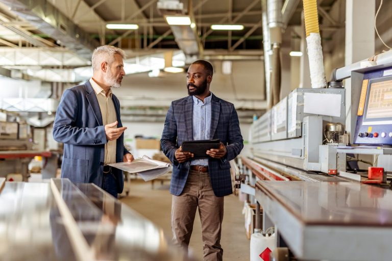 Two men in blazers talking in a factory beside industrial machinery, one holding a tablet and the other papers