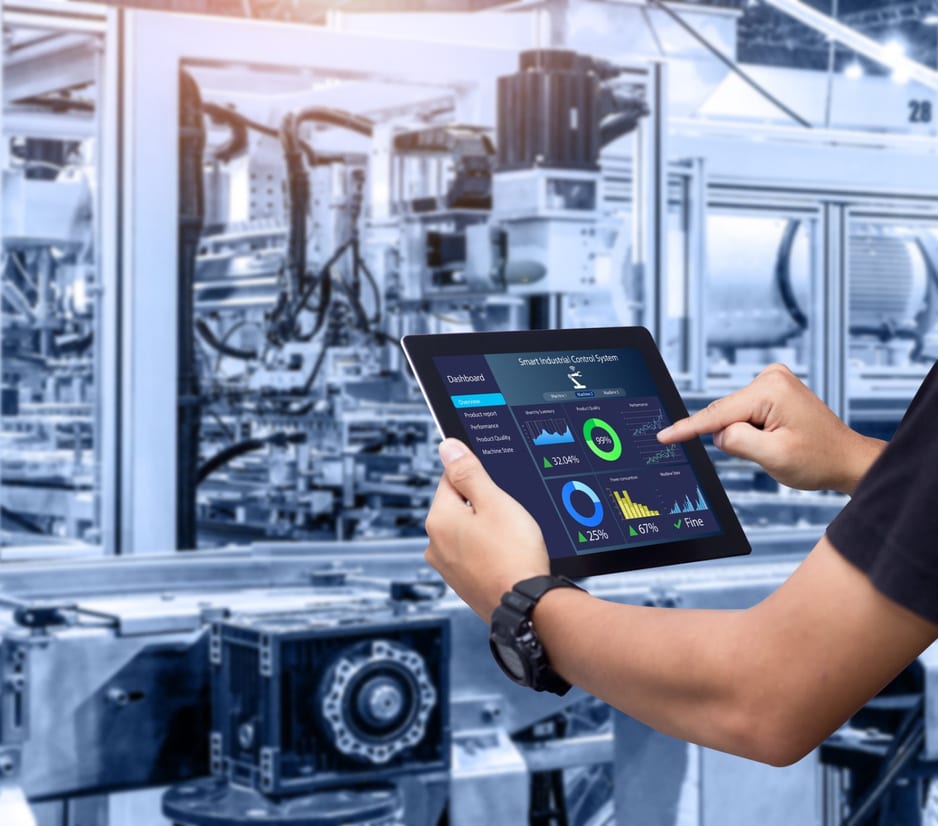 Person holding a tablet showing an industrial control dashboard with charts, in front of automated factory machinery