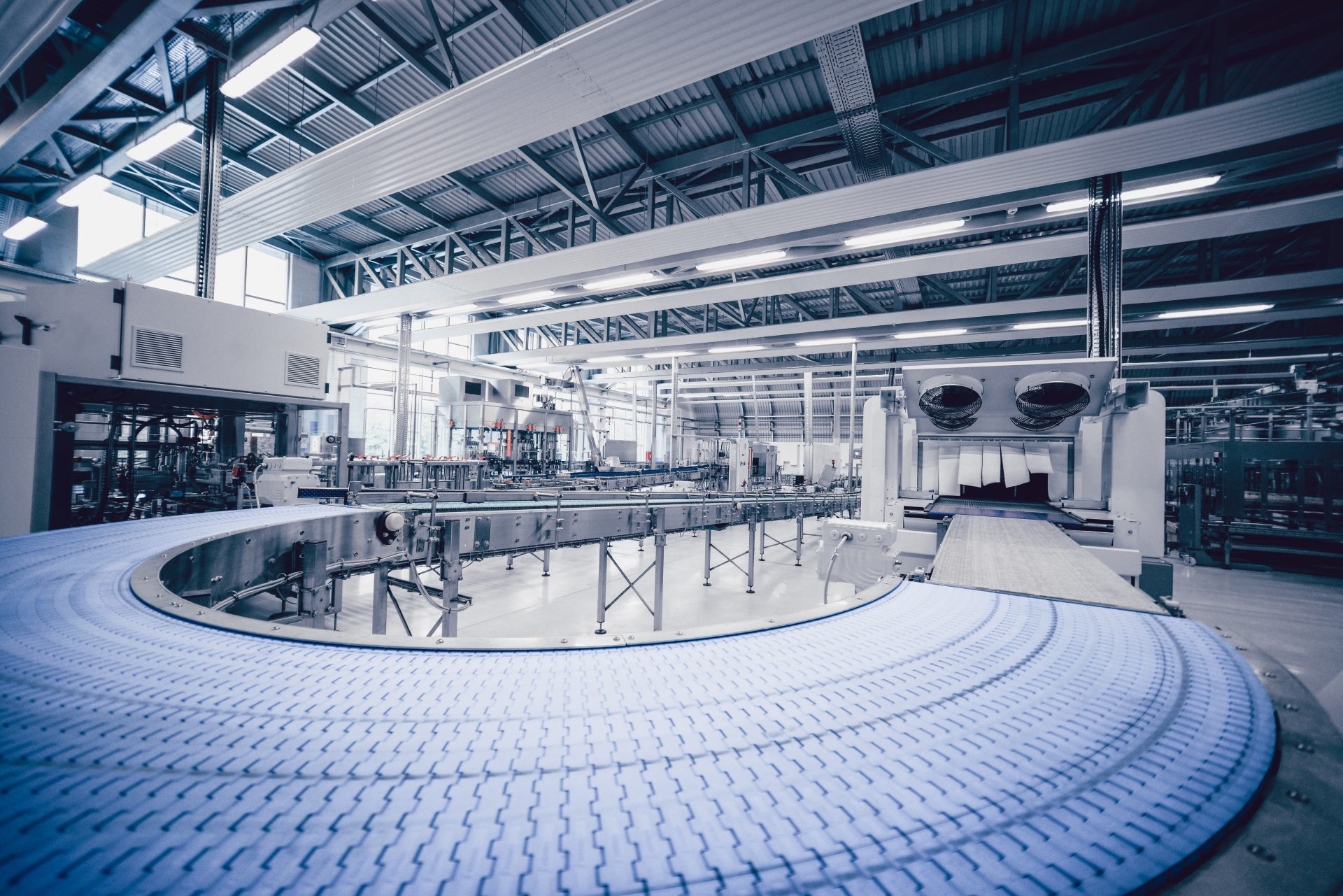 Curved conveyor belt in a large, empty factory hall with steel roof trusses and production machinery in the background
