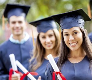 Five smiling graduates in dark gowns and mortarboards holding rolled certificates tied with red ribbon outdoors