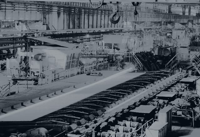 Interior of a steelworks with a long steel strip on a roller conveyor beneath an overhead crane hook