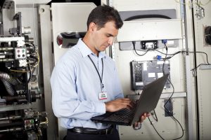 Maintenance engineer in a blue shirt and lanyard using a laptop beside industrial machinery and a control cabinet