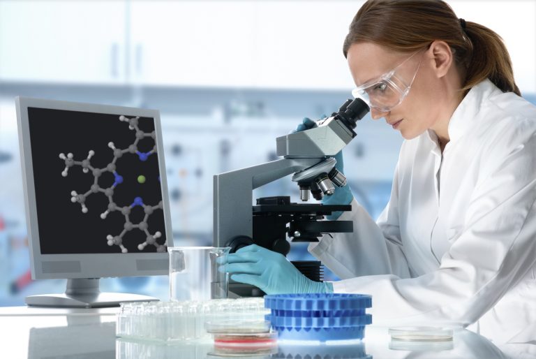 Scientist in a white lab coat, goggles and gloves looks into a microscope beside a screen showing a molecular model