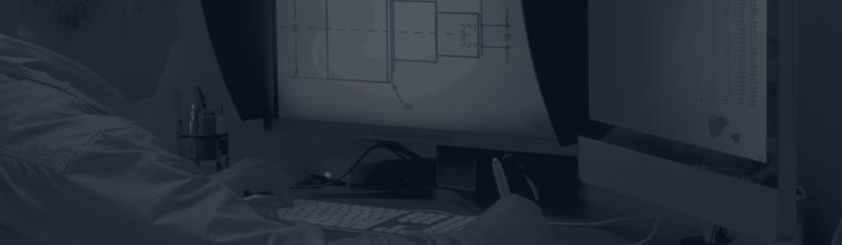 Engineer at a desk looking at a technical drawing of a machined part on a computer monitor, with a keyboard in front