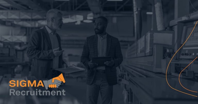Two men in blazers talking in a factory beside industrial machinery, one holding a tablet