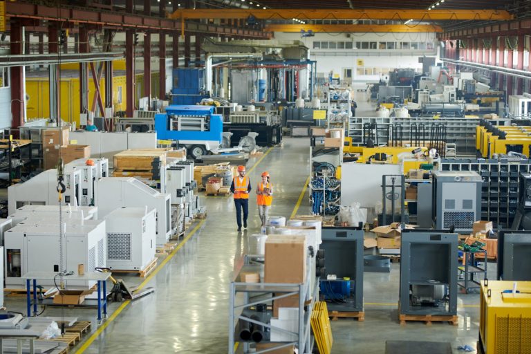 Two workers in hi-vis vests and hard hats walking through a large factory floor with machinery and crates